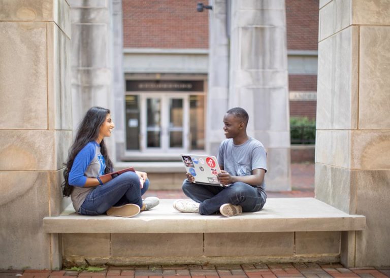 Two students on UK's campus working on a computer together. One has long, black wavy hair and is wearing a UK t-shirt and jeans. The other has short, black curly hair and is wearing a grey t-shirt.