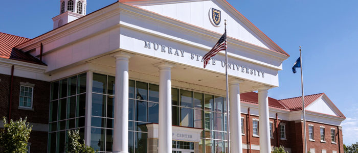 Murray State University's welcome center. It is a building with four pillars and a bell tower. Out front, the American and Kentucky flag are flying. The sky is clear and blue.