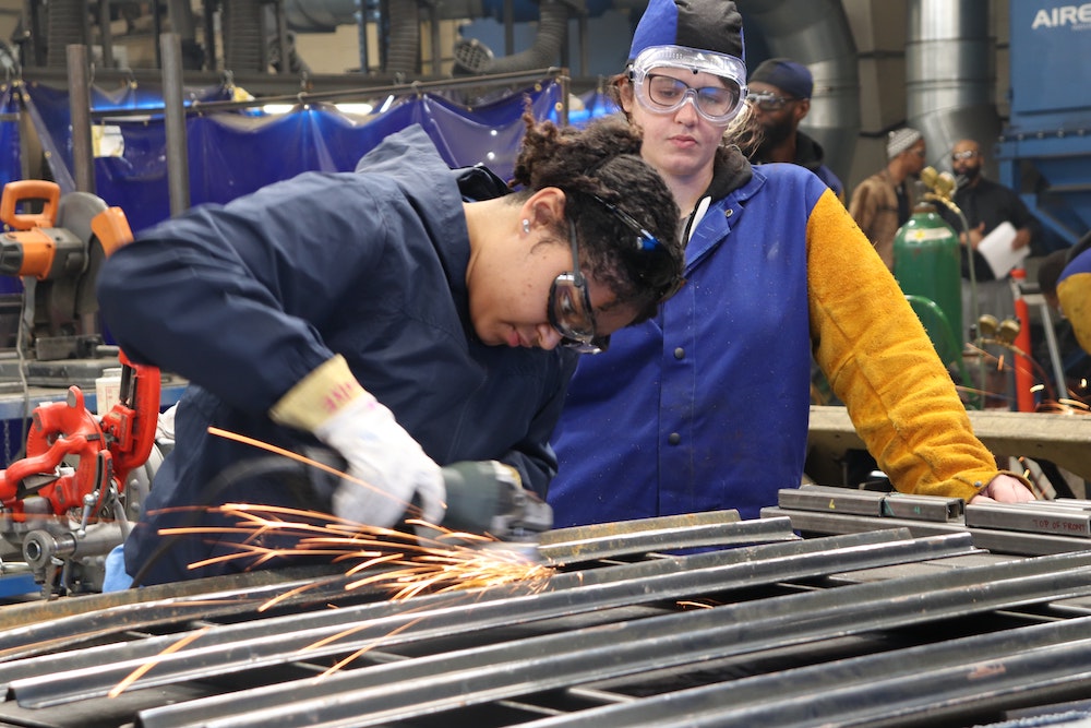 Two women welding. They are both wearing safety goggles and gloves.