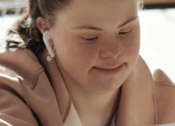 Woman with Down Syndrome wearing headphones and smiling at her computer. She is wearing a pink jacket and has her brown hair tied in a pony tail.