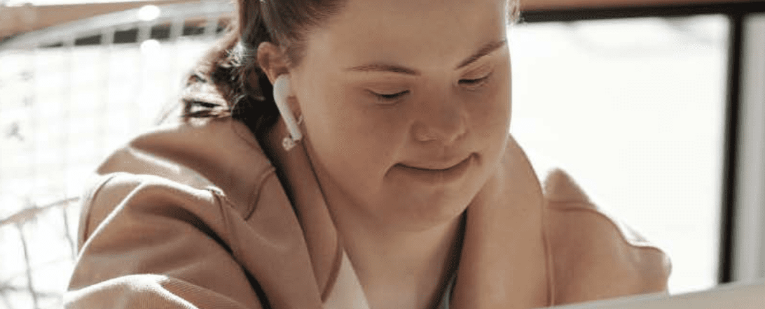 Woman with Down Syndrome wearing headphones and smiling at her computer. She is wearing a pink jacket and has her brown hair tied in a pony tail.