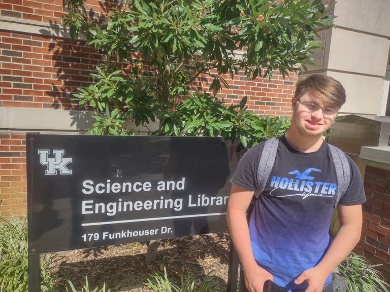 A college student stands in front of a building sign at the University of Kentucky campus.