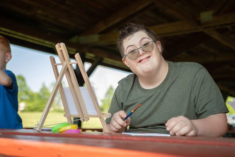 A person is painting in a park shelter. On the table before them are a canvas and paints.